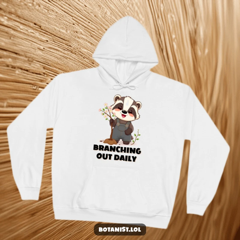 Funny Badger Hoodie depicting a happy badger in overalls, humorously pruning a small, blooming tree, offering warmth and a smile.